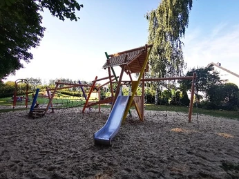Kombispielgerät Ein Spielplatz mit Klettergerüst und Rutsche auf sandigem Boden, umgeben von Bäumen und Grünflächen.