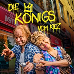 Zwei fröhliche Personen posieren lächelnd vor einem Kiosk mit buntem Graffiti-Schriftzug.