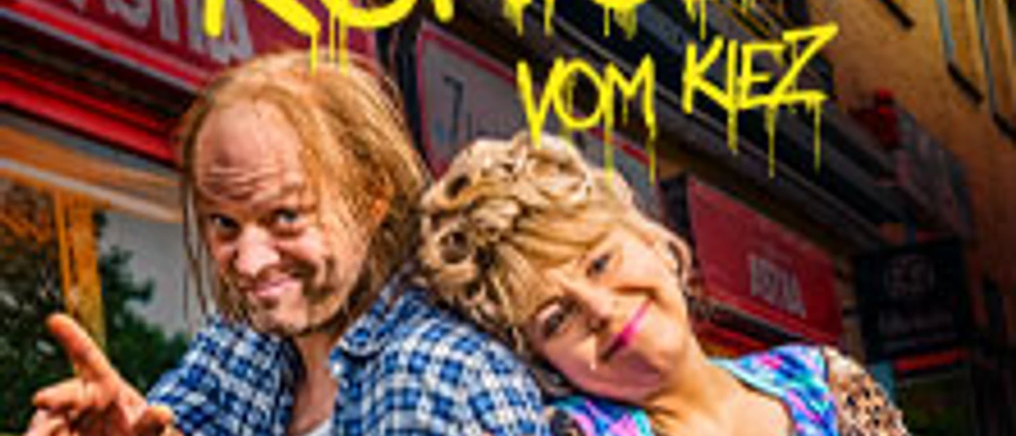 Zwei fröhliche Personen posieren lächelnd vor einem Kiosk mit buntem Graffiti-Schriftzug.