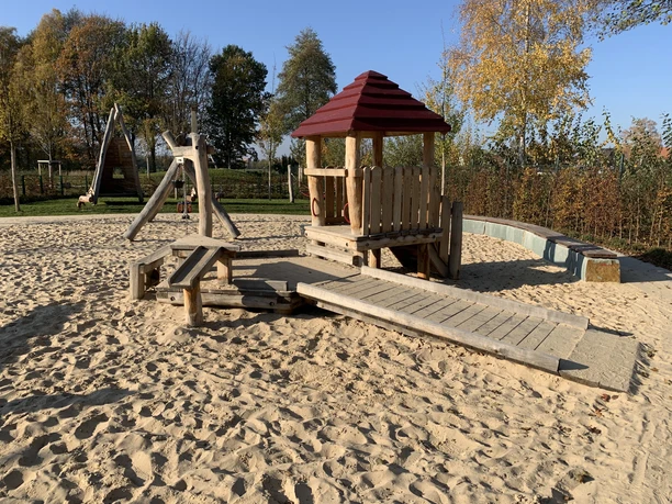 Holzspielplatz mit Rutsche und Sandkasten, umgeben von Bäumen, an einem sonnigen Herbsttag.