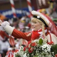 Funkemariechen Rote Funken Eine junge Frau in traditionellem rot-weißen Kostüm lächelt und hat Blumen schmückend in der Hand.A young women in a traditional red and white costume smiles and holds flowers in her hand.