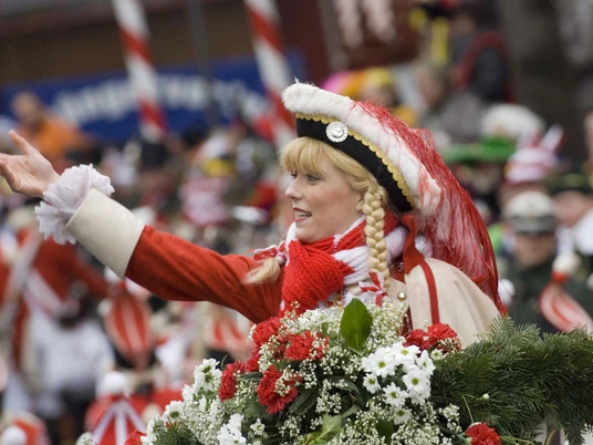 Funkemariechen Rote Funken Eine junge Frau in traditionellem rot-weißen Kostüm lächelt und hat Blumen schmückend in der Hand.A young women in a traditional red and white costume smiles and holds flowers in her hand.