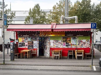 Kebapland Blick auf das Imbisshaus. Die Fassade ist rot gestrichen, hat eine rote Markise und Sitzmöglichkeiten davor.View of the snack bar. The façade is painted red, has a red sunshade, and tables and chairs in front.