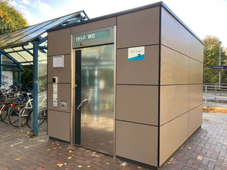 Bahnhof Ratzeburg_WC1_Petra Roemer_20241022.jpg Am Bahnhof steht ein öffentliches Toilettenhäuschen mit silberner Tür, daneben überdachte Fahrradstellplätze
