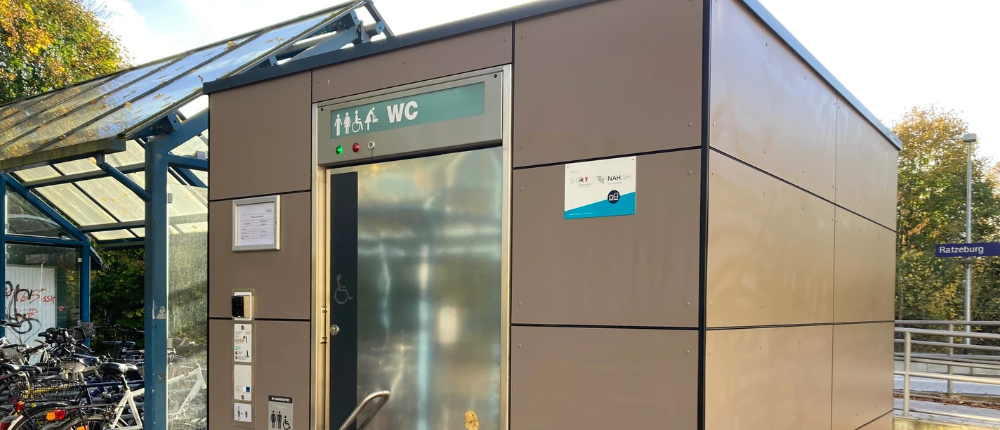 Bahnhof Ratzeburg_WC1_Petra Roemer_20241022.jpg Am Bahnhof steht ein öffentliches Toilettenhäuschen mit silberner Tür, daneben überdachte Fahrradstellplätze