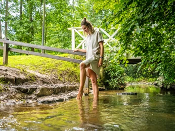 Frau genießt einen Spaziergang im Wasser auf dem Barfußpfad, umgeben von üppigem Grün im Kurpark.