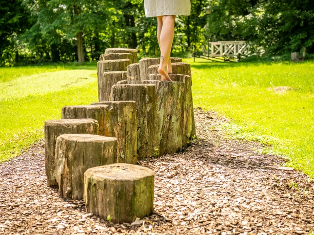 Person geht barfuß über Holzstämme im Barfußpfad im Kurpark Bad Holzhausen, umgeben von Natur.