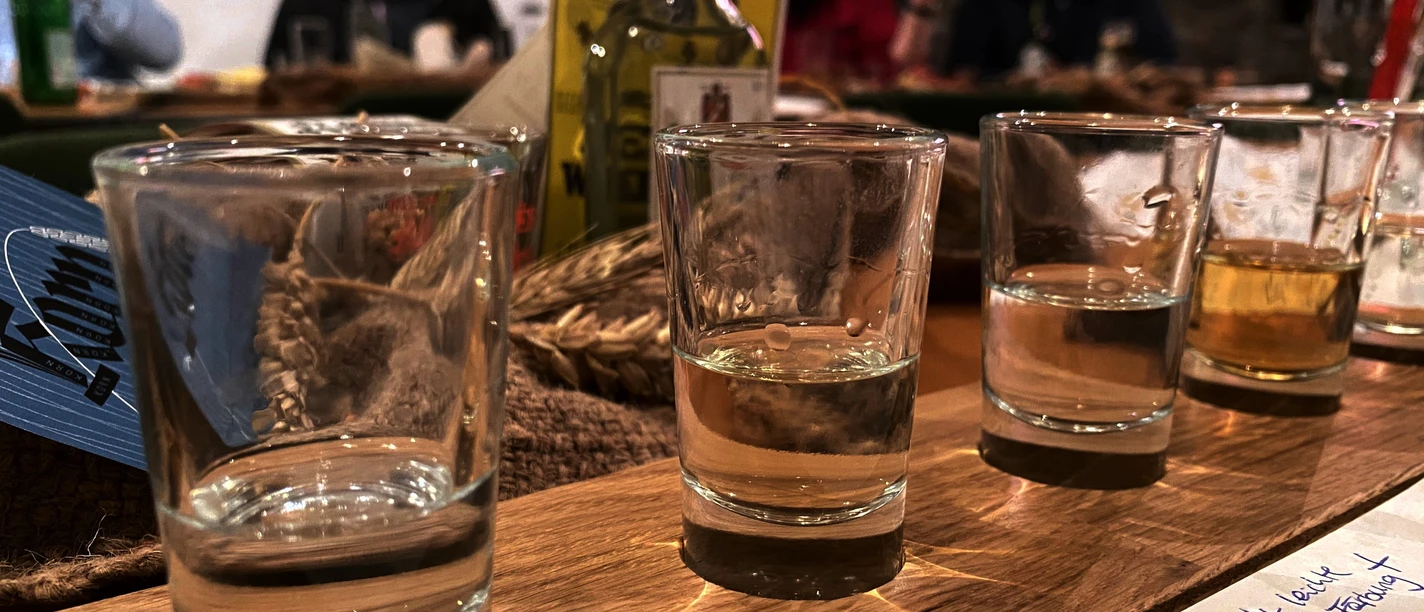 Alte Brennerei Mehrere Gläser mit klarer Flüssigkeit stehen auf einem Holztisch, im Hintergrund unscharfe Personen.