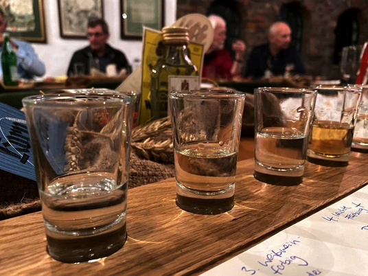 Alte Brennerei Mehrere Gläser mit klarer Flüssigkeit stehen auf einem Holztisch, im Hintergrund unscharfe Personen.