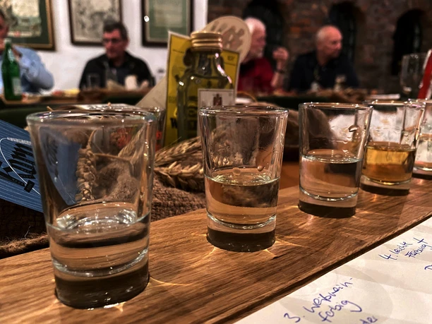 Alte Brennerei Mehrere Gläser mit klarer Flüssigkeit stehen auf einem Holztisch, im Hintergrund unscharfe Personen.