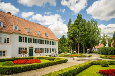 Ein historisches Gebäude mit rotem Dach vor einer gepflegten Gartenanlage und einem runden Brunnen.
