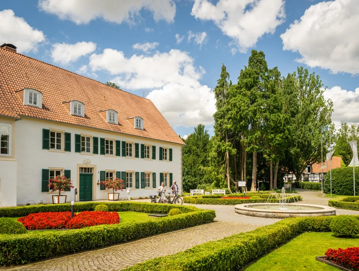 Kurpark Bad Holzhausen Ein historisches Gebäude mit rotem Dach vor einer gepflegten Gartenanlage und einem runden Brunnen.