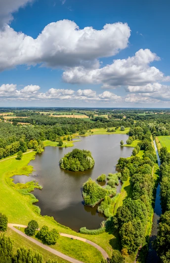 Luftaufnahme eines ruhigen Sees mit umliegenden grünen Wiesen und dichten Wäldern an einem sonnigen Tag.