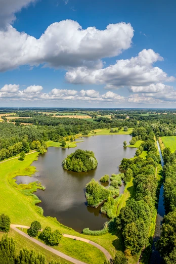Luftaufnahme eines ruhigen Sees mit umliegenden grünen Wiesen und dichten Wäldern an einem sonnigen Tag.