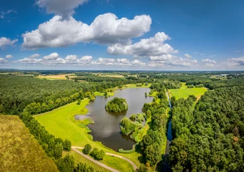 Kleihügelsee Luftaufnahme eines ruhigen Sees mit umliegenden grünen Wiesen und dichten Wäldern an einem sonnigen Tag.