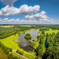 Kleihügelsee Luftaufnahme eines ruhigen Sees mit umliegenden grünen Wiesen und dichten Wäldern an einem sonnigen Tag.