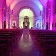 Kirchenschiff beleuchtet in violettem und blauem Licht, mit Holzbänken und einem Altar im Hintergrund.