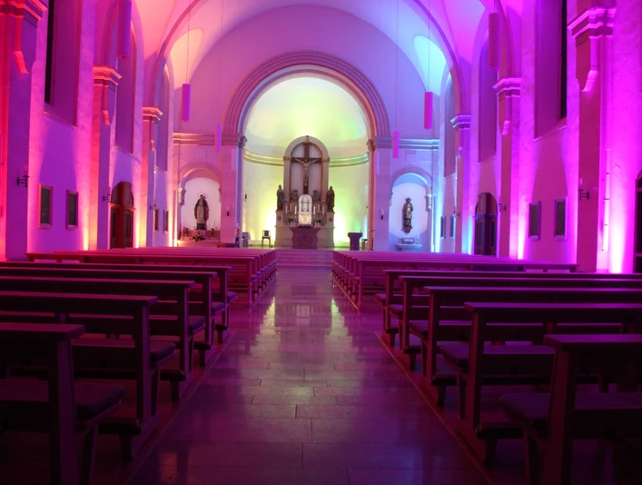 LichterHimmel-Kirche Westerwiehe (1).JPG Kirchenschiff beleuchtet in violettem und blauem Licht, mit Holzbänken und einem Altar im Hintergrund.