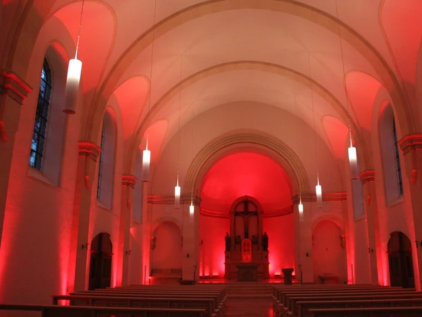 Innenansicht einer Kirche im roten Licht, mit gewölbter Decke, hängenden Leuchten und Altar.