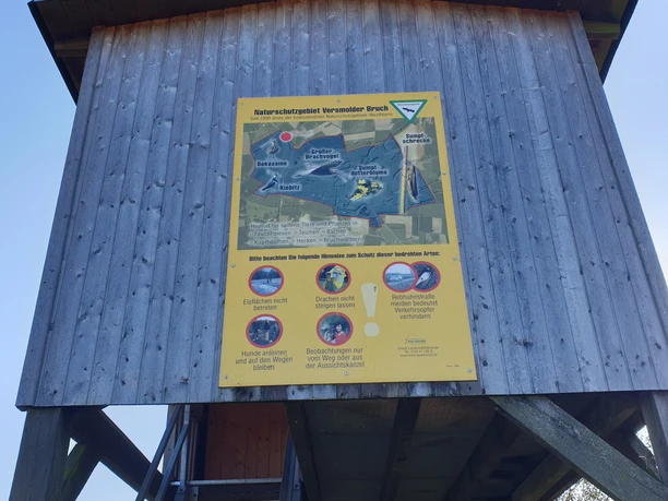 Holzturm mit gelbem Infoplakat im Naturschutzgebiet. Erkundungsstation für Besucher und Naturfreunde.