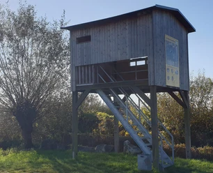 Vogelbeobachtungsplateau.jpg Eine erhöhte Vogelbeobachtungsplattform aus Holz mit Treppe und Sichtfenster in einer ländlichen Landschaft.
