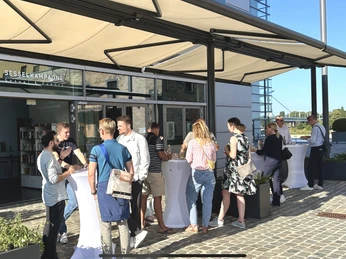 Hafenterrasse mit Sonnenmarkise Menschen treffen sich bei Sonnenschein auf der Terrasse mit weißen Stehtischen und SonnensegelPeople meet in the sunshine on the terrace with white bar tables and awning