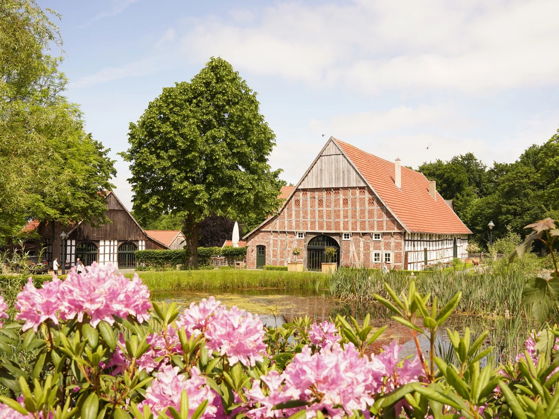 Landluftkonzert_Fotocredit-NWD.jpg Fachwerkhäuser mit rotem Giebel, umgeben von blühenden Rhododendren und einem kleinen Teich.