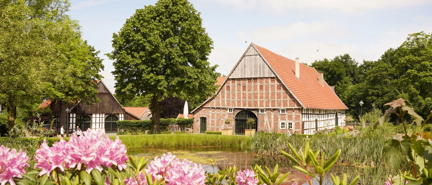 Landluftkonzert_Fotocredit-NWD.jpg Fachwerkhäuser mit rotem Giebel, umgeben von blühenden Rhododendren und einem kleinen Teich.
