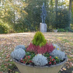 Ein herbstlicher Park mit einer dekorativen Pflanzschale im Vordergrund und einem Kunstwerk im Hintergrund.