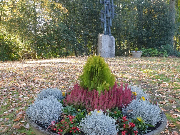 Ein herbstlicher Park mit einer dekorativen Pflanzschale im Vordergrund und einem Kunstwerk im Hintergrund.