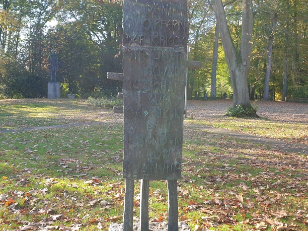 Gedenktafel aus Holz im herbstlichen Park, umgeben von Laub und Bäumen, erinnert an Kriegsopfer.