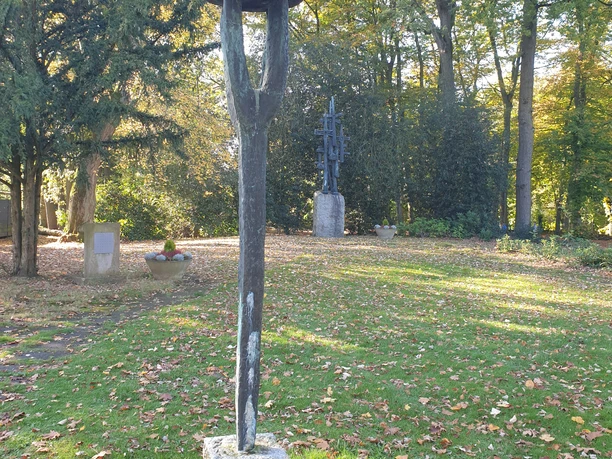Skulptur in einem parkähnlichen Friedhof mit Rasen, Laub und Bäumen bei Sonnenlicht.
