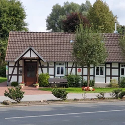 Ein Fachwerkhaus mit rotem Ziegeldach, grünem Vorgarten und umliegenden Bäumen an einer Straße.