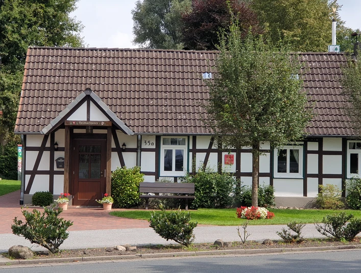Ein Fachwerkhaus mit rotem Ziegeldach, grünem Vorgarten und umliegenden Bäumen an einer Straße.