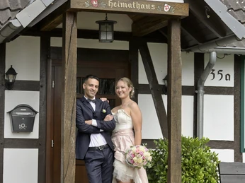 Ein Brautpaar posiert lächelnd vor einem traditionellen Fachwerkhaus, geschmückt mit Blumen.