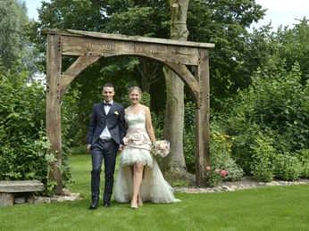Standesamt Versmold_0731_01.JPG Ein Brautpaar spaziert glücklich durch einen gepflegten, grünen Garten unter einem rustikalen Holzbogen.
