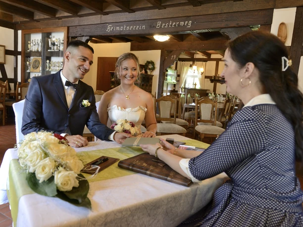Ein junges Hochzeitspaar sitzt in einem rustikalen Gastraum gegenüber einer Standesbeamtin.