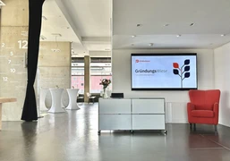 Köln.Business Wirtschaftsförderung X Sessel Hub Rheinauhafen Registrierungscounter mit Display im Hintergrund und Blick in den Konferenzbereich mit StehtischenRegistration counter with display in the background and view of the conference area with bar tables