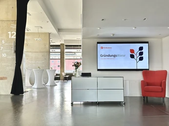 Köln.Business Wirtschaftsförderung X Sessel Hub Rheinauhafen Registrierungscounter mit Display im Hintergrund und Blick in den Konferenzbereich mit StehtischenRegistration counter with display in the background and view of the conference area with bar tables