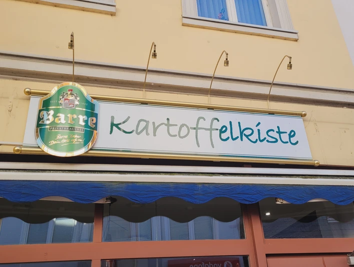 Außenansicht eines Gebäudes mit einem Schild der Gaststätte "Kartoffelkiste" und einem Barre-Bierschild.
