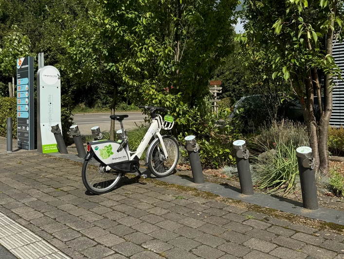 E-Bike Verleihstation Weiße Fahrräder an einer grauen Leihstation, umgeben von Bäumen und einem Weg im Hintergrund.