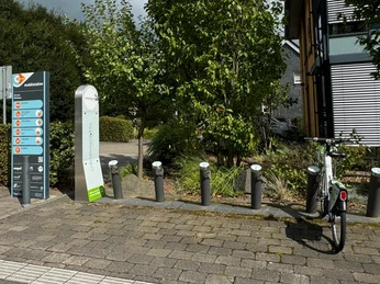 E-Bike Verleihstation Fahrradparkplätze mit Ladesäulen, ein Fahrrad steht bereit, umgeben von Bäumen und einem modernen Gebäude.