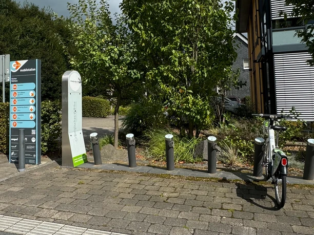 E-Bike Verleihstation Fahrradparkplätze mit Ladesäulen, ein Fahrrad steht bereit, umgeben von Bäumen und einem modernen Gebäude.