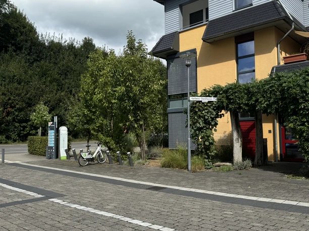 E-Bike Verleihstation Gelbes Gebäude mit modernem Design, umgeben von Bäumen und Fahrradbügel. Straßenzeichen sichtbar.