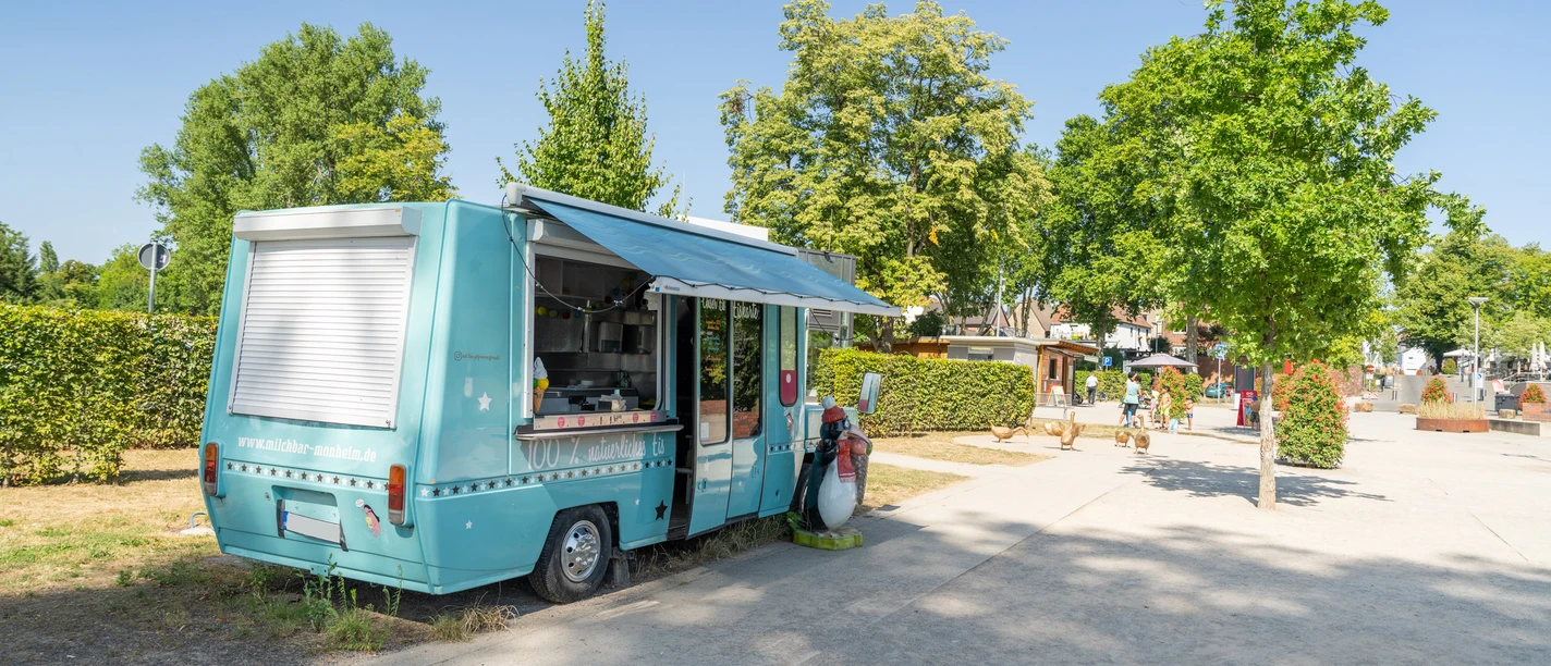 Eiswagen Fred Ein türkisfarbener Eiswagen steht auf einem Gehweg, umgeben von grünen Bäumen und Sträuchern.