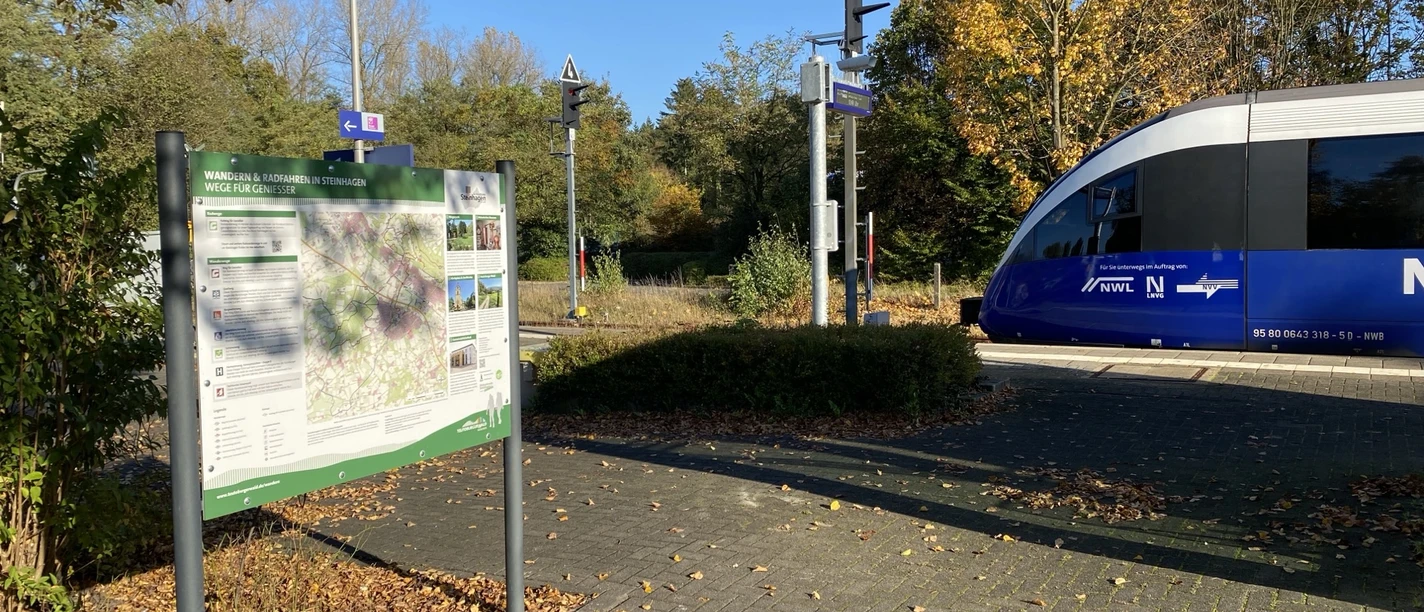 Infotafel Bahnhof Steinhagen Bahnübergang im Herbst, links Infotafel, rechts blauer Zug, umgeben von goldenen Herbstbäumen.