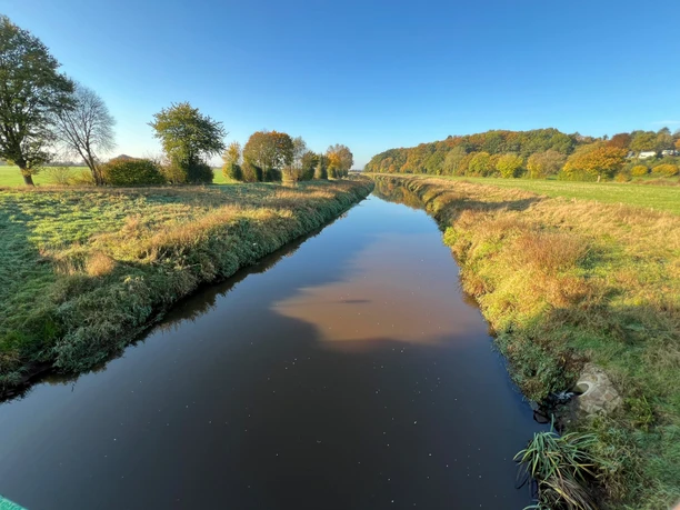 Ein ruhiger Fluss fließt durch eine herbstliche Landschaft mit Bäumen und einem klaren blauen Himmel.