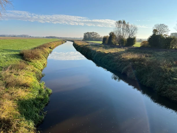 Ein stiller Fluss fließt durch eine flache Landschaft, gesäumt von Bäumen und unter einem klaren Himmel.