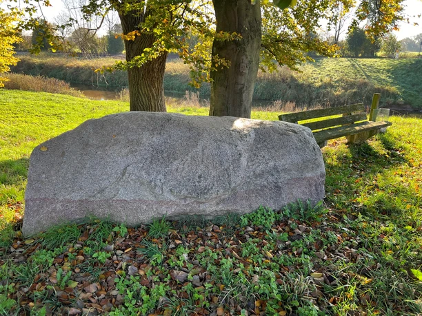 A large boulder in a meadow near two trees, flanked by a simple wooden bench in the fall.