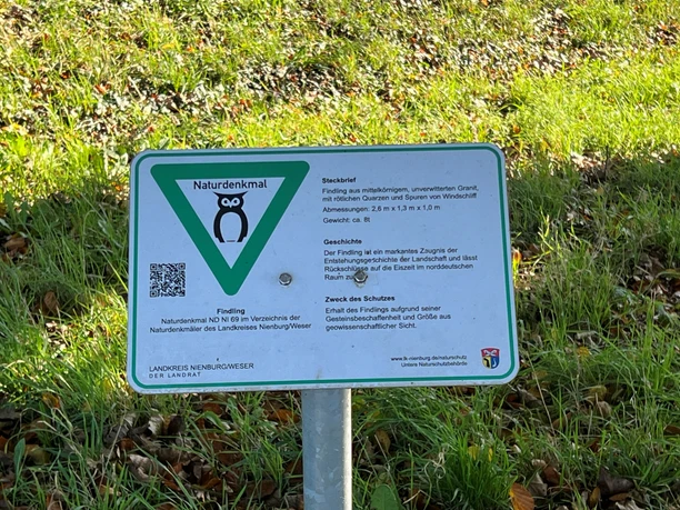 A large boulder stands as a natural landmark in a meadow, flanked by an information sign.